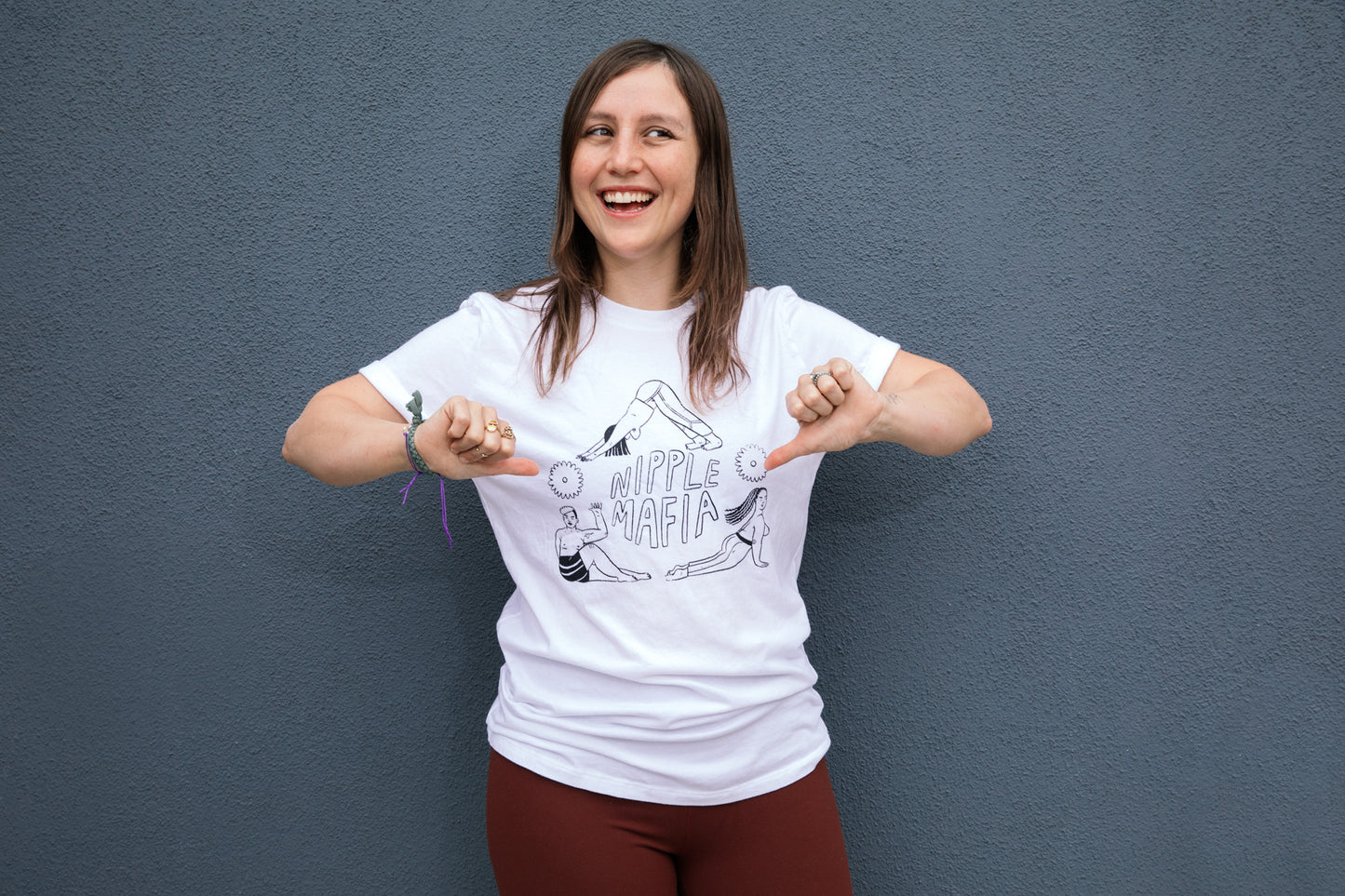 person pointing with their thumbs to the white tee shirt their wearing that says NIPPLE MAFIA written in black surrounded by three people in different yoga poses that form a triangle around the print
