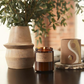 amber glass jar with lit candle on a table with a plant and a ceramic pot